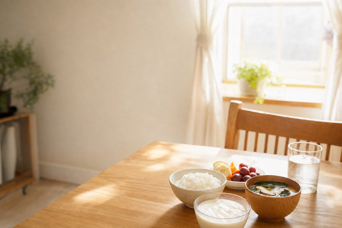 朝の自然光が差し込むキッチンのテーブルに、ヨーグルト、ごはんとみそ汁、果物、水の入ったグラスが並び、窓の外に朝の光が見える