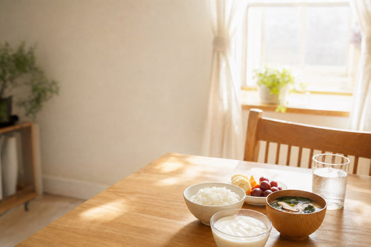 朝の自然光が差し込むキッチンのテーブルに、ヨーグルト、ごはんとみそ汁、果物、水の入ったグラスが並び、窓の外に朝の光が見える