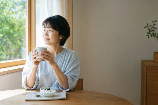 朝の光が差し込む窓辺で、お茶のカップを両手で包み、穏やかに外を眺める女性。テーブルにはブルーベリーをのせたヨーグルトの小鉢。腸と脳のつながりをテーマにした記事のアイキャッチ