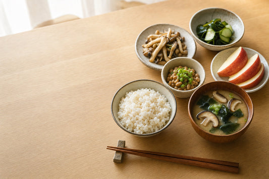 朝の自然光が差し込む木のテーブルに、麦ごはん、具だくさんのみそ汁、納豆、わかめの酢の物、きのこのソテー、くし切りのりんごが並んだ和の朝食